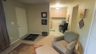 This single-floor residence offers a well-organized and functional layout designed for comfortable living. The entryway greets visitors with a tiled foyer and a welcome mat, opening into a carpeted living area that provides a warm and relaxed atmosphere. The living space is accented by decorative wall-mounted frames, adding a personal touch to the neutral, earthy-toned walls. A plush armchair with an ottoman anchors the seating area, complemented by a floral area rug that adds color and texture to the space. Adjacent to the living area, the kitchen and dining space feature light wood cabinetry, offering ample storage, and tile flooring that transitions seamlessly from the entryway. The dining area accommodates a round table with seating, making it an ideal spot for meals or casual gatherings. A small potted plant near the dining area introduces a touch of greenery, enhancing the overall warmth of the space. The textured ceiling and soft, ambient lighting throughout contribute to the cozy and inviting character of this well-appointed single-floor home.