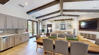 This single-story community clubhouse combines rustic architectural charm with modern amenities in a well-appointed, open-concept layout. The interior is immediately distinguished by soaring vaulted ceilings adorned with dark, exposed wood beams that run the length of the space, creating a warm and inviting atmosphere. Large-format tile flooring in a neutral tone flows consistently throughout, unifying each functional area.

The main gathering area seamlessly integrates a dining zone and lounge space. A substantial wood dining table surrounded by upholstered gray chairs sits beneath a striking rectangular chandelier, making it well-suited for group gatherings. The adjacent lounge features a generous sectional sofa accented with patterned throw pillows, woven ottomans, and a large wall-mounted flat-screen television set within a wood-paneled surround against a textured accent wall.

The kitchen is fully equipped with stainless steel appliances, including a gas range, refrigerator, and dishwasher, complemented by gray shaker-style cabinetry, white countertops, and a mosaic tile backsplash. Multiple sets of glass French doors connect the interior to an expansive outdoor area featuring a large swimming pool, lounge seating, and mature palm trees, allowing abundant natural light to fill the space.