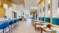 This expansive single-floor amenity and clubhouse space offers a thoughtfully designed interior that blends coastal-modern aesthetics with functional versatility. The open layout flows seamlessly across distinct zones, including a welcoming reception and leasing area, a social lounge, a coffee and refreshment bar, and access to a fitness center.

The design palette centers on crisp white walls accented by bold teal blue and warm gold tones, creating a lively yet sophisticated atmosphere. Oversized woven rattan pendant lights hang dramatically from the high ceilings, complemented by starburst wall sconces and layered recessed lighting that collectively create a warm, inviting ambiance throughout.

Seating options are abundant and varied — from velvet teal barstools lining a sleek white counter to curved wood lounge chairs paired with marble-top accent tables, and teal upholstered banquette booths with warm wood framing. The lounge area features distinctive pod-style seating and a communal table, ideal for gatherings or focused work.

Large windows and glass doors allow generous natural light to filter through, enhanced by flowing patterned drapery. Decorative laser-cut wood panels and abstract artwork add artistic detail throughout. Light-toned tile flooring runs consistently across the space, grounding the design. A textured yellow accent wall behind the coffee bar adds a playful focal point, making this a dynamic and welcoming community hub.