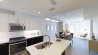 This contemporary single-floor residence showcases a thoughtfully designed open-concept layout that seamlessly connects the kitchen, dining, and living areas. The kitchen is a standout feature, boasting a large central island with a crisp white quartz countertop and a double stainless steel sink topped with a sleek pull-down faucet. A striking set of pendant lights hangs above the island, adding both elegance and focused illumination. The cabinetry features a bold two-tone design, pairing crisp white upper cabinets with dark lower cabinets, complemented by a classic white subway tile backsplash. Stainless steel appliances, including a built-in microwave and range, complete the modern culinary space. The open flow leads into a bright dining area, where natural light pours in through large windows, illuminating the warm hardwood-style flooring that runs throughout. Recessed lighting and clean white walls enhance the airy, spacious ambiance across the entire space. The overall aesthetic is polished and modern, offering a welcoming and functional environment ideal for both everyday living and entertaining.