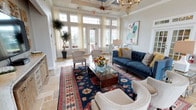 This beautifully appointed single-floor space makes an immediate impression with its refined architectural details and sophisticated interior design. The centerpiece of the space is a grand lounge area that showcases soaring coffered ceilings, adorned with a decorative ceiling fan and an ornate crystal chandelier that together create a sense of grandeur and elegance.

Tall, multi-pane transom windows and classic French doors flood the interior with generous natural light, framing outdoor views and lending the space an airy, open atmosphere. Sheer ivory curtains soften the light and complement the neutral wall tones throughout.

The lounge is thoughtfully furnished with a deep navy tufted sofa, paired with neutral accent chairs and a glass-topped coffee table, creating a balanced and inviting seating arrangement. A richly patterned area rug in navy, red, and cream anchors the space, adding warmth and visual interest to the light stone tile flooring beneath.

Curated decorative touches — including a vibrant floral arrangement, colorful accent pillows, a large abstract artwork, and a dramatic indoor palm — bring personality and life to the room. The overall ambiance is one of polished comfort, blending classic architectural character with eclectic, colorful furnishings to create a welcoming and visually dynamic environment.