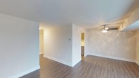 This single-floor space offers a clean, modern aesthetic with an open and airy layout. The living area is finished with durable wood-style plank flooring that runs throughout, providing a warm and cohesive look. Neutral white walls and smooth ceilings keep the space feeling bright and welcoming, while a ceiling fan with an integrated light fixture adds both style and practical comfort to the main living area. The open floor plan allows for a natural flow between the living space and the kitchen, which is accessible through a wide doorway. The kitchen features warm wood-toned cabinetry and appears well-appointed for everyday use. A partial wall divider subtly separates the main living zone from an adjoining area, creating a sense of definition without sacrificing openness. Recessed lighting accents the hallway area, contributing to the overall well-lit ambiance of the unit. Clean white baseboards throughout complement the light color palette and add a polished finishing touch. The overall atmosphere is fresh, move-in ready, and well-suited for a range of furnishing styles, making it a versatile and functional living space.