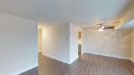 This single-floor space offers a clean, modern aesthetic with an open and airy layout. The living area is finished with durable wood-style plank flooring that runs throughout, providing a warm and cohesive look. Neutral white walls and smooth ceilings keep the space feeling bright and welcoming, while a ceiling fan with an integrated light fixture adds both style and practical comfort to the main living area. The open floor plan allows for a natural flow between the living space and the kitchen, which is accessible through a wide doorway. The kitchen features warm wood-toned cabinetry and appears well-appointed for everyday use. A partial wall divider subtly separates the main living zone from an adjoining area, creating a sense of definition without sacrificing openness. Recessed lighting accents the hallway area, contributing to the overall well-lit ambiance of the unit. Clean white baseboards throughout complement the light color palette and add a polished finishing touch. The overall atmosphere is fresh, move-in ready, and well-suited for a range of furnishing styles, making it a versatile and functional living space.