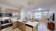 This single-floor residence offers a thoughtfully designed open-concept layout that seamlessly connects the kitchen, dining, and living areas. The kitchen is a standout feature, showcasing taupe cabinetry, quartz countertops, a large peninsula with seating, stainless steel appliances including a French door refrigerator and electric range, and elegant glass pendant lighting overhead. Light wood-style flooring runs throughout the main living areas, enhancing the bright and airy atmosphere created by multiple large windows dressed with sheer curtains and blinds.

The living room is a welcoming space with natural light pouring in through a front-facing window alongside a classic paneled entry door. Two well-proportioned bedrooms each feature ceiling fans, generous windows with outdoor views of green surroundings, and light wood flooring. A hallway with a closet connects the private areas of the home.

The bathroom is finished with marble-look tile flooring, a tiled walk-in shower, a single-sink vanity with cabinetry, and a well-lit mirror. An additional flex room serves as a functional home office, complete with large windows, ample natural light, and generous space for a full desk setup and shelving. The overall ambiance is clean, contemporary, and inviting throughout.
