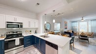 This single-floor, 2-bedroom, 2-bathroom residence is designed with a modern aesthetic and practical layout that maximizes comfort and flow. The open-concept kitchen and living area serves as the heart of the home, featuring a striking two-tone kitchen with bold navy lower cabinets, crisp white upper cabinetry, and a classic subway tile backsplash. A spacious quartz peninsula with a stainless steel sink provides ample prep space and casual seating, while pendant lighting and stainless steel appliances complete the polished look. The living room receives abundant natural light through large windows dressed with sheer curtains and blinds, and features crown molding and a ceiling fan overhead. Both bedrooms are carpeted and well-sized, with the primary bedroom offering generous proportions, crown molding, and a walk-in closet with wire shelving for organized storage. The second bedroom also benefits from natural light and comfortable proportions. The bathrooms are finished in clean white tones with modern vanities, large mirrors, and a shower-tub combination. Two hallways connect the spaces efficiently, and two closets provide additional storage throughout. Light wood-look flooring runs through the main living areas, tying the space together with a warm, contemporary feel.