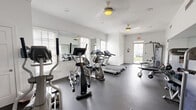 Located at 108 Lords Court in Virginia Beach, VA, this fitness center offers a well-equipped workout space designed to support a comprehensive exercise routine. The single-room facility is thoughtfully laid out to maximize functionality within its compact footprint.

The space features durable rubber flooring throughout, providing excellent traction and cushioning for varied workout activities. Bright white walls, crown molding, and wainscoting lend the room a clean, polished aesthetic. Ceiling fans help maintain comfortable airflow during exercise sessions, while large windows fitted with plantation shutters allow natural light to fill the space.

Cardio equipment includes multiple treadmills and elliptical machines, along with a stationary bike, catering to a range of cardiovascular training needs. A multi-function cable machine and a weight bench with free weights — including a well-organized dumbbell rack — provide ample options for strength training. A wall-mounted flat-screen television adds entertainment during workouts, and a water cooler station ensures easy hydration. Large wall mirrors enhance the sense of openness while allowing users to monitor their form.

A French-style glass-panel door leads to an adjacent area, and a secondary exit with a glass door opens to the outdoors, contributing to the room's bright and airy ambiance. Posted fitness center rules and informational signage round out this practical, inviting community amenity.