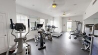 Located at 108 Lords Court in Virginia Beach, VA, this fitness center offers a well-equipped workout space designed to support a comprehensive exercise routine. The single-room facility is thoughtfully laid out to maximize functionality within its compact footprint.

The space features durable rubber flooring throughout, providing excellent traction and cushioning for varied workout activities. Bright white walls, crown molding, and wainscoting lend the room a clean, polished aesthetic. Ceiling fans help maintain comfortable airflow during exercise sessions, while large windows fitted with plantation shutters allow natural light to fill the space.

Cardio equipment includes multiple treadmills and elliptical machines, along with a stationary bike, catering to a range of cardiovascular training needs. A multi-function cable machine and a weight bench with free weights — including a well-organized dumbbell rack — provide ample options for strength training. A wall-mounted flat-screen television adds entertainment during workouts, and a water cooler station ensures easy hydration. Large wall mirrors enhance the sense of openness while allowing users to monitor their form.

A French-style glass-panel door leads to an adjacent area, and a secondary exit with a glass door opens to the outdoors, contributing to the room's bright and airy ambiance. Posted fitness center rules and informational signage round out this practical, inviting community amenity.