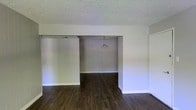 This single-story space presents a straightforward, functional layout characterized by clean lines and a neutral color palette throughout. The living area flows openly into an adjoining dining space through a wide structural opening, creating a connected and cohesive floor plan well-suited for everyday living and entertaining.

The flooring throughout is a rich, dark hardwood-style material that provides a grounding contrast to the light gray and white walls. White baseboards frame the perimeter of each room, adding a crisp, finished look. The textured popcorn ceiling adds a classic touch overhead, while a simple pendant light fixture hangs centrally in the dining area, providing focused overhead illumination.

The open passage between the two rooms enhances the sense of depth and space, allowing light and sightlines to travel freely. The walls are freshly painted in a soft, neutral tone, offering a versatile backdrop for a variety of interior design styles. A door visible on the right side of the living area suggests additional rooms or utility spaces beyond. Overall, the space offers a clean, move-in-ready environment with a practical and unpretentious aesthetic.