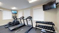 Located at 1655 Gilmore Park Ave in Columbus, Ohio, this fitness center offers a clean, well-organized workout environment designed to support a variety of exercise routines. The single-floor facility is thoughtfully laid out to maximize usability and comfort for its users.

The workout area is equipped with multiple treadmills and an elliptical machine, providing ample options for cardiovascular training. A dedicated free weights section features a fully stocked dumbbell rack with a wide range of weight options, catering to both beginner and experienced fitness enthusiasts. Rubber flooring throughout the workout space provides safety and stability during exercise.

Two wall-mounted flat-screen televisions are positioned for easy viewing from the cardio equipment, allowing users to stay entertained during workouts. The space is well-lit with overhead fluorescent lighting, ensuring a bright and functional environment at any time of day.

Natural light is introduced through multiple windows fitted with white horizontal blinds, contributing to the open and airy feel of the room. The neutral color palette of the walls and clean aesthetic of the space create a focused, distraction-free atmosphere conducive to an effective workout. Overall, this fitness center provides a practical and inviting setting for daily exercise.