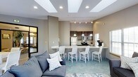 This single-floor space showcases an open-concept layout that connects the living area and kitchen in a cohesive, functional design. The living area is anchored by a comfortable gray sectional sofa accented with decorative pillows, set atop a patterned area rug. The herringbone-laid teal tile flooring adds a distinctive design element that runs throughout the shared space, contributing to its contemporary character.

The kitchen is equipped with stainless steel appliances, including a refrigerator, range, and microwave, complemented by dark cabinetry and a light-toned countertop. A spacious breakfast bar extends from the kitchen island, lined with white bar stools that seat multiple guests — ideal for casual dining or entertaining.

Overhead, a geometric coffered ceiling with recessed lighting creates visual interest while brightening the space. Track lighting along the kitchen ceiling provides additional task illumination. Pendant lights hang above the bar area, adding warmth and dimension. A large mirror near the living area enhances the sense of openness. Neutral warm-gray walls tie the entire space together with a clean, polished aesthetic.