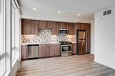 Located at 17600 Cartwright Rd in Irvine, CA, this single-floor unit presents a clean, modern aesthetic throughout. Light wood-tone flooring flows seamlessly across the entire space, creating a cohesive and airy feel. The kitchen is a standout feature, outfitted with rich wood-grain cabinetry extending to the ceiling, a striking mosaic tile backsplash in neutral tones, white quartz countertops, and a full suite of stainless steel appliances including a gas range, over-the-range microwave, refrigerator, and dishwasher. The layout also includes a dedicated sink area with ample counter space and natural light from an adjacent window. The main living area is bright and open, anchored by a wide bank of floor-to-ceiling windows fitted with roller shades, offering views of palm trees and the surrounding community. The bathroom is well-appointed with large-format white wall tiles, a glass-enclosed walk-in shower, and warm wood accent details. The hallway connects the spaces efficiently and includes recessed lighting and a utility panel. Throughout, the unit maintains crisp white walls, recessed lighting, and a minimalist design sensibility that reflects contemporary Irvine living.