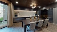 This elegantly designed single-floor space blends upscale aesthetics with versatile functionality. The centerpiece is a generous dining area anchored by a substantial dark wood table surrounded by tufted velvet chairs in soft grey and blue tones. Overhead, an ornate embossed metallic ceiling tile in deep bronze commands attention, complemented by a sculptural globe chandelier that casts warm, ambient light throughout the room. A full wet bar and kitchenette runs along one wall, featuring backlit mosaic tile, dark cabinetry, stainless steel appliances, and a marble-style countertop — ideal for entertaining. Gold candle holders serve as a refined centerpiece detail on the dining table. Adjacent to the main dining area, a dramatic private booth lounge features wrap-around gold velvet banquette seating, dark lacquered walls, recessed lighting, and marble-topped cocktail tables, creating an intimate, speakeasy-inspired atmosphere. A separate seating nook with deep blue accent chairs and framed artwork opens to the outdoors through floor-to-ceiling sliding glass windows draped with sheer linen curtains, allowing natural light and views of lush exterior landscaping. Wide-plank hardwood flooring flows cohesively throughout, tying together the distinct zones of this polished, hospitality-forward space.