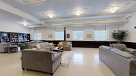 Located at 751 2nd Ave S in Clinton, Iowa, this single-floor space offers a generous and well-appointed layout designed for both relaxation and gathering. The standout living room commands attention with its ornate coffered ceiling featuring decorative molding and geometric detailing — a hallmark of classic architectural craftsmanship. Dark wainscoting lines the lower walls throughout, creating a striking two-tone aesthetic that pairs elegantly with the bright white upper walls. Multiple windows dressed with roller and horizontal blinds allow ample natural light to fill the space. The living area is furnished with multiple seating arrangements, including plush sofas and accent chairs centered around coffee tables, making it ideal for social interaction. A built-in bookcase filled with books adds a warm, library-like ambiance to one corner, complemented by a wall-mounted television nearby. The dining room serves a dual purpose as a meeting space, featuring a large conference-style table surrounded by ergonomic chairs, and is decorated with playful card suit artwork. Additional café-style seating offers a more casual setting. The light-toned flooring throughout ties the spaces together, creating a cohesive and welcoming environment well-suited for community use.