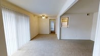 This single-floor residence offers a straightforward, functional layout with a comfortable living area as its central feature. The living space is finished with neutral wall-to-wall carpeting and light-toned walls, creating a clean, understated atmosphere throughout. A ceiling fan with a built-in light fixture is centrally mounted, providing both air circulation and overhead lighting to the room.

Vertical blinds line the window along one wall, offering adjustable light control and a degree of privacy. The open layout connects seamlessly to adjacent areas, with a hallway visible in the background leading to additional rooms, suggesting a practical flow between living and private spaces.

A notable architectural detail is the pass-through opening in the wall that connects the living area to the kitchen. This feature allows visibility into the kitchen, which is equipped with wood-toned cabinetry and a white refrigerator, promoting a sense of openness between the two spaces. The overall ambiance is modest and move-in ready, with neutral finishes throughout that provide a versatile backdrop suitable for a variety of interior styles.