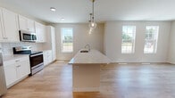 This well-designed single-floor residence offers a clean, modern aesthetic throughout, with light-toned finishes and an efficient open layout that seamlessly connects the kitchen, dining, and living areas. The kitchen is a standout feature, equipped with white shaker-style cabinetry, light granite countertops, a classic white subway tile backsplash, and a full suite of stainless steel appliances including a refrigerator, electric range, and over-the-range microwave. A generously sized center island with an undermount sink and elegant pendant lighting serves as both a functional workspace and a social hub. Light hardwood-style flooring flows throughout the main living areas, contributing to the bright, airy atmosphere enhanced by multiple windows that invite ample natural light. The living area extends toward sliding glass doors that open to a private balcony, offering pleasant outdoor views. The bedroom features soft carpet flooring and a closet, with a clear sightline to the kitchen beyond. A dedicated laundry area houses a stacked full-size washer and dryer unit alongside built-in wire shelving for added storage. The bathroom is finished with neutral tile flooring, a white vanity with mirror, and accessibility-friendly grab bars, rounding out this thoughtfully appointed space.