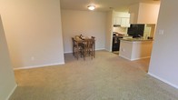 This single-floor residence offers a practical and comfortable open-concept layout designed to maximize usable living space. The main living and dining area is finished with wall-to-wall neutral carpeting, lending a warm and cozy atmosphere throughout. A dining nook sits naturally within the open floor plan, providing a defined space for meals without feeling enclosed. The kitchen is positioned adjacent to the dining area and is separated by a peninsula counter, offering a natural division between cooking and living spaces while maintaining an open feel. The kitchen is equipped with black appliances, including a range and refrigerator, and features white cabinetry with overhead storage. Dark countertops complement the cabinetry, while a tiled floor grounds the kitchen zone distinctly from the carpeted living areas. Recessed flush-mount ceiling lighting illuminates the main living space, contributing to a clean and understated aesthetic. Neutral-toned walls throughout create a versatile backdrop, making the space easy to personalize. The overall design is straightforward and functional, making this a comfortable single-level living environment suited for everyday use.