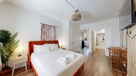 Located at 805 Fourth Ave N in the heart of Saint Petersburg, Florida, this efficiently designed one-bedroom, one-bathroom unit offers a thoughtfully arranged single-floor layout. The bedroom serves as the centerpiece of the space, featuring warm wood-look flooring, a bold orange-upholstered bed frame, and patterned curtains that add a pop of color against crisp white walls. A distinctive slatted wood accent wall houses a wall-mounted flat-screen TV, adding texture and visual interest, while a small wooden table and chair set provides a cozy nook. A woven pendant light fixture completes the inviting ambiance. The compact kitchen is efficiently outfitted with stainless steel appliances, including an electric range, white cabinetry with matte black hardware, a single-basin sink with a black gooseneck faucet, and a tile backsplash — everything needed for convenient meal preparation. The bathroom is designed with accessibility in mind, featuring grab bars, a roll-in style shower with a curtain enclosure and rainfall showerhead, and wood-look flooring that ties the space together cohesively. A wall-mounted wooden shelf provides practical storage. The overall aesthetic is clean, modern, and welcoming, with a warm color palette throughout.