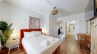 Located at 805 Fourth Ave N in the heart of Saint Petersburg, Florida, this efficiently designed one-bedroom, one-bathroom unit offers a thoughtfully arranged single-floor layout. The bedroom serves as the centerpiece of the space, featuring warm wood-look flooring, a bold orange-upholstered bed frame, and patterned curtains that add a pop of color against crisp white walls. A distinctive slatted wood accent wall houses a wall-mounted flat-screen TV, adding texture and visual interest, while a small wooden table and chair set provides a cozy nook. A woven pendant light fixture completes the inviting ambiance. The compact kitchen is efficiently outfitted with stainless steel appliances, including an electric range, white cabinetry with matte black hardware, a single-basin sink with a black gooseneck faucet, and a tile backsplash — everything needed for convenient meal preparation. The bathroom is designed with accessibility in mind, featuring grab bars, a roll-in style shower with a curtain enclosure and rainfall showerhead, and wood-look flooring that ties the space together cohesively. A wall-mounted wooden shelf provides practical storage. The overall aesthetic is clean, modern, and welcoming, with a warm color palette throughout.