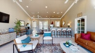 This single-floor clubhouse is designed to serve as a welcoming community hub, blending comfort and functionality across several distinct areas. The main lounge is generously proportioned, featuring soaring ceilings with crown molding, recessed lighting, and ceiling fans that contribute to an airy, open atmosphere. Light tile flooring, layered area rugs, and a cohesive coastal-inspired palette of white, teal, and warm neutrals unify the space. Plush seating arrangements — including sofas, accent chairs, and coffee tables — invite relaxed gatherings, while large windows with white shutters allow abundant natural light to fill the room. A bar-height counter with mid-century modern stools provides an additional social focal point. Adjacent to the main lounge is a cozy dining nook with a round glass-top table, complemented by direct outdoor access through a glass-paned door. A fully equipped kitchen area features granite countertops, gray cabinetry, a stainless steel range, and a microwave, making it well-suited for events and catering needs. A dedicated private office, distinguished by a bold red accent wall and warm hardwood-style flooring, offers a functional workspace complete with a ceiling fan and ample room for consultations. Throughout the clubhouse, thoughtful design details and quality finishes create a polished, inviting environment.