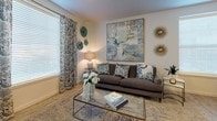 This single-floor residence offers a thoughtfully designed layout that balances comfort and functionality across its key living spaces. The bedroom is a serene retreat, featuring plush carpeting, a tufted upholstered bed frame, mirrored nightstands, and large windows dressed with elegant patterned curtains and blinds that allow natural light to filter in while maintaining privacy. Framed wall art adds a refined finishing touch to the space. The living area exudes a warm, curated ambiance with light wood-style flooring, a large statement painting as a focal point, and stylish décor including sunburst wall accents and patterned drapes. A glass-top coffee table and a plush sofa complete this inviting gathering space. The kitchen is well-equipped with crisp white shaker cabinetry, dark granite countertops, and a full suite of stainless steel appliances including a range, over-the-range microwave, dishwasher, and refrigerator. Light wood-style flooring flows throughout, adding warmth and continuity. The bathroom features a clean, functional design with white cabinetry, a dark granite vanity top, wood-look tile flooring, a full soaking tub with shower, and a bright vanity mirror with overhead lighting. This space offers a cohesive and comfortable living environment throughout.