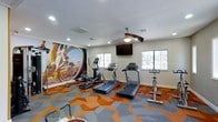 This fitness center offers a focused and energizing environment for residents to pursue their wellness goals. The space is anchored by a striking full-wall mural depicting a cyclist in action, setting an athletic tone throughout the room. The vibrant geometric-patterned carpet in shades of orange and gray adds a modern, dynamic energy to the floor, complementing the room's active purpose.

The cardio suite is well-appointed, featuring multiple treadmills, an elliptical trainer, and a stationary spin bike, providing a range of options for cardiovascular exercise. A wall-mounted flat-screen television is positioned for easy viewing during workouts, while a ceiling fan with a light fixture ensures comfortable air circulation throughout the session.

Natural light filters in through multiple windows along the walls, creating a bright and welcoming atmosphere. Recessed lighting supplements the natural light, ensuring the space remains well-lit at all hours. The room's layout is thoughtfully organized, allowing ample space between machines for safe and comfortable use. The clean, white walls and updated fixtures give the fitness center a fresh, modern appearance that encourages an active and healthy lifestyle.