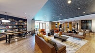 This expansive clubroom is thoughtfully designed across a single floor, offering a diverse range of social and recreational spaces united by a cohesive contemporary aesthetic. The main lounge area serves as the centerpiece, featuring tufted leather sofas, accent chairs in rich cognac tones, and a patterned area rug atop warm hardwood floors. A striking teal-finished textured ceiling panel defines this central gathering space, lending visual depth and a boutique-hotel atmosphere.

A dedicated library nook provides floor-to-ceiling bookshelves stocked with reading material, complemented by work tables and chairs—ideal for quiet study or casual work. Adjacent to this is a fireplace lounge with a wall-mounted television and stacked wood display, creating an inviting, intimate retreat.

The games zone features a billiards table, mounted flat-screen TV, and a built-in bar counter with stools, finished with dark accent walls and large-format tile flooring. Reclaimed wood wall paneling appears throughout, adding warmth and texture across zones.

Large glass doors and floor-to-ceiling windows provide abundant natural light and direct outdoor access to a courtyard pool area. Decorative laser-cut metal artwork and curated furnishings complete the polished, urban design.
