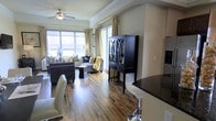 This thoughtfully designed single-floor residence offers an open-concept layout that seamlessly connects the living and kitchen areas, creating a spacious and inviting atmosphere. Rich, warm-toned hardwood flooring runs throughout, adding character and continuity to the space.

The living area is generously sized and bathed in natural light, courtesy of large floor-to-ceiling windows dressed with elegant gold and ivory curtains. A ceiling fan with a classic light fixture anchors the room overhead, while crown molding lends a refined architectural touch. The space is furnished with plush seating, layered rugs, and accent pieces in a cohesive palette of charcoal, gold, and cream, creating a sophisticated yet comfortable ambiance. A striking black display cabinet with decorative glass panels serves as a standout focal point, adding a touch of drama to the room.

The kitchen area flows naturally from the living space, featuring dark granite countertops and a breakfast bar with modern bar seating. Built-in appliances and rich cabinetry complete the culinary space with a sleek, functional aesthetic.

Sliding glass doors provide access to an outdoor area, allowing natural light to pour in and enhancing the overall sense of openness. The entire residence strikes an ideal balance between elegance and livability.