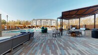 Located at 2421 Landmark Dr in Raleigh, NC, this outdoor amenity space at Leigh House is designed to deliver a resort-style experience for its residents. The centerpiece of the area is a generously sized swimming pool, lined with lounge chairs and umbrella-shaded seating that invite relaxation under open skies. A broad, multi-toned paver courtyard extends throughout the space, providing a polished and cohesive aesthetic that ties all areas together. Decorative teal ceramic urns serve as striking focal points along the poolside promenade.

A covered pavilion with exposed brick construction anchors one end of the amenity area, housing a comfortable lounge with plush seating, a wall-mounted television, and an outdoor fireplace — creating an inviting year-round gathering spot. Large glass-paneled doors open into the adjacent interior amenity facility, branded with the Leigh House logo, which appears to include fitness equipment and additional services. A separate pergola-covered zone features built-in grilling stations, dining tables, and lush landscaping, offering an ideal setting for outdoor cooking and entertaining. Privacy fencing with warm wood tones frames the perimeter of the space, enhancing seclusion while complementing the brick and paver materials throughout. The overall ambiance is upscale, cohesive, and thoughtfully designed for communal outdoor living.