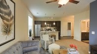 This single-story residence showcases an open-concept design that connects the living area and kitchen in one cohesive, flowing space. The vaulted ceiling adds a sense of height and airiness, complemented by a ceiling fan with wooden blades that enhances both comfort and style. Large windows along the walls invite abundant natural light, brightening the warm, neutral-toned interior.

The living area features plush carpeting layered with a patterned area rug, providing a comfortable and inviting atmosphere. Tasteful seating arrangements, including tufted and upholstered accent chairs, contribute to the space's welcoming character.

The kitchen is outfitted with dark espresso cabinetry contrasted by light countertops, stainless steel appliances—including a refrigerator, microwave, and range—and pendant lighting that hangs above the kitchen island or bar-height counter. Track lighting overhead ensures functional illumination across the workspace.

A hallway visible beyond the kitchen leads to additional areas of the home, suggesting a well-organized and practical floor plan. Overall, the space balances modern aesthetics with everyday functionality in a bright, open setting.