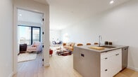 This single-floor residence is designed with a contemporary aesthetic, combining clean lines, neutral tones, and thoughtfully selected materials throughout. The open-concept layout connects the kitchen, dining, and living areas, creating a cohesive and functional living space filled with natural light.

The kitchen is well-appointed with stainless steel appliances, including a range, microwave, and refrigerator. Light wood upper cabinetry contrasts with crisp white lower cabinets, and a marble-style backsplash adds visual interest. A substantial concrete-finish island with bar seating serves as both a workspace and gathering point, seamlessly bridging the kitchen and living area.

The living and dining zone is bright and open, featuring recessed lighting, white walls, and light hardwood flooring that runs throughout the unit. Large windows allow natural light to flow into the space, contributing to an airy, welcoming ambiance.

The bedroom is calm and well-proportioned, with a window offering outdoor views and ample space for furnishings. Two bathrooms are finished with stone-tile flooring and warm wood vanities on black metal frames. One bathroom includes a glass-enclosed shower, while the other features a soaking tub, both maintaining a consistent, modern design language.