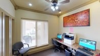 The business center is a single-floor workspace designed to meet essential professional needs. The room features a granite-topped workstation that accommodates a desktop computer and a multifunction printer, making it suitable for tasks such as printing, scanning, and copying. A ceiling fan with integrated lighting provides both air circulation and illumination, complementing the recessed lighting fixtures installed throughout. Natural light filters into the space through a large window fitted with horizontal blinds, offering a view of the exterior surroundings while allowing light control. The walls are finished in a warm neutral tone, creating a calm and focused atmosphere. A vibrant abstract canvas in rich reds and oranges adds a decorative accent, bringing visual interest to the workspace. Carpeted flooring and ergonomic task chairs contribute to a comfortable working environment. A waste bin is conveniently positioned near the workstation. The overall layout is efficient and well-organized, providing residents or guests with a dedicated, fully equipped space to handle business or personal tasks with ease.