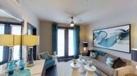 This single-floor space showcases a thoughtfully designed living area centered around a harmonious teal and neutral color scheme. The room is anchored by a ceiling fan with a built-in light fixture, ensuring both comfort and ambiance. Large French doors serve as a focal point, flooding the space with natural light while offering views to the outdoors, framed by rich teal drapes that add depth and elegance.

The living area is furnished with a plush gray sofa accented with teal and patterned throw pillows, complemented by a cozy accent chair in matching teal upholstery. A set of modern white circular side tables provides functional surface space with a contemporary aesthetic. The light-toned area rug grounds the seating arrangement and adds texture underfoot.

Wall decor includes a striking large-scale abstract artwork in shades of blue and white, reinforcing the coastal-inspired color palette. Warm table lamps on either side of the room provide layered lighting. The neutral wall tones and white ceiling create a clean, open backdrop that ties all design elements together into a cohesive and inviting living environment.