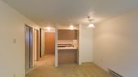 This single-floor space showcases an open-concept layout that seamlessly connects the living area to the kitchen. The kitchen is defined by warm wood cabinetry that extends to the upper walls, providing ample storage. A peninsula-style counter with a pass-through design creates a natural division between the kitchen and living space while maintaining an open, airy feel. A decorative pendant light hangs above the counter area, adding a focal point to the space. The flooring throughout the living area features neutral-toned carpet, contributing to a warm and comfortable atmosphere. A textured ceiling runs throughout, consistent with the overall design aesthetic. A hallway is visible toward the back of the space, leading to additional rooms and a solid wood entry door, suggesting thoughtful layout and privacy. Recessed and surface-mounted lighting provides even illumination throughout. The overall color palette — combining beige tones, warm wood finishes, and white walls — creates a cohesive and inviting environment suited for everyday living.