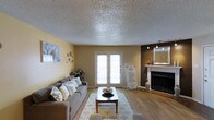 Located in Lancaster, this single-floor space offers a thoughtfully designed interior that balances comfort and style. The living area serves as the heart of the space, featuring warm hardwood-style flooring that runs throughout and a textured ceiling that adds visual depth. A classic white fireplace with a decorative mantle anchors one side of the room, creating a cozy and inviting focal point. On the opposite wall, a bold, dark accent wall adds contrast and sophistication, complemented by track lighting and a large framed mirror that enhances the sense of openness. Natural light pours generously into the room through a set of French doors, which provide access to an outdoor area and lend an airy, bright atmosphere to the space. The living area is spacious enough to accommodate generous seating arrangements, and the neutral yellow-toned walls provide a warm, welcoming backdrop. Decorative touches throughout — including wall art, accent vases, and a patterned area rug in soft blues and yellows — contribute to an overall ambiance that feels both curated and comfortable. This space is well-suited for everyday living and entertaining alike.