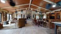 This inviting clubhouse offers a generously sized, single-floor layout designed for both comfort and communal gathering. The centerpiece of the space is a dramatic vaulted ceiling with exposed white-painted wooden beams and rich wood paneling, lending a warm, lodge-like atmosphere throughout. Large format tile flooring flows seamlessly across the open-plan living area, anchored by a striking full-height brick fireplace adorned with an oversized decorative clock, creating a bold focal point. Multiple seating arrangements — including wicker chairs, upholstered sofas, and ornate wooden tables — define distinct conversation and relaxation zones within the expansive great room. Natural light filters in generously through numerous windows dressed with bamboo shades and patterned drapes, while multiple ceiling fans ensure comfortable airflow. A dedicated dining nook with a round table and cross-back chairs offers an intimate setting alongside the main living area. The kitchen features a granite-topped island with bar seating, stainless steel appliances, dark wood cabinetry, and distinctive woven pendant lighting. A hallway connects the main spaces and provides a graceful transition through the clubhouse. Throughout, the décor strikes a balance between rustic charm and refined character, making this clubhouse a welcoming and functional gathering destination.