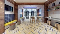 This thoughtfully designed lobby makes a strong first impression through its layered mix of materials, color, and functional furnishings. The eye-catching herringbone-patterned floor, rendered in tones of cream, yellow, and grey, anchors the entire space and flows seamlessly throughout. Warm wood paneling lines the walls and ceiling in key zones, adding a natural richness that balances the bold use of cobalt blue paint found near the elevator alcove.

The entrance features a full-height glazed storefront with black-framed glass doors, allowing natural light to pour in and providing clear views to the street beyond. Adjacent to the entry, a built-in bench upholstered in plaid fabric with a bright yellow seat cushion offers a casual waiting area, accented by dual wall-mounted pendant lights above. Compact round stools nearby provide additional informal seating.

A communal high-top table with bar-height chairs sits centrally, encouraging interaction and providing a practical surface for residents or visitors. A concierge or package management station is positioned along one wall, adding a functional service element to the space.

The elevator vestibule is framed by a graceful arch, with stainless steel elevator doors set against a clean light grey backdrop, all anchored by the same distinctive patterned flooring. The overall ambiance is energetic, welcoming, and contemporary.
