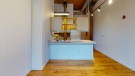 Located at 51 Front St in Cumberland, Rhode Island, this single-floor space is a striking example of industrial mill conversion architecture. The unit masterfully balances historic character with modern functionality throughout its open layout.

The main living area commands immediate attention with its soaring ceilings featuring original wood beam and plank construction, exposed silver ductwork, and rich red brick walls. Expansive arched multi-pane windows flood the space with abundant natural light while framing views of the surrounding landscape, lending the room a gallery-like openness. Original hardwood floors run throughout, adding warmth and continuity.

The kitchen area is efficiently designed, featuring a wraparound peninsula with a light-toned countertop and integrated sink, open shelving with warm wood accents, and overhead task lighting. A separate galley-style kitchen section offers ample cabinetry in a bold golden-yellow finish, a stainless steel sink, and a full suite of appliances including a range and microwave, all set atop durable tile flooring.

The bathroom is finished in clean, classic white tile surrounding a full soaking tub and shower combination, complemented by recessed lighting, a large vanity mirror, and warm wood cabinetry with a speckled countertop — providing a crisp, functional retreat within the industrial aesthetic of the broader space.