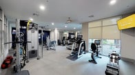 This spacious fitness center is designed to accommodate a full range of workout needs within a clean, modern environment. The facility is organized into distinct training zones, beginning with an extensive cardio area housing multiple Life Fitness treadmills, elliptical trainers, and stair climbers arranged along floor-to-ceiling windows. The windows are fitted with roller shades and offer views of outdoor greenery, allowing natural light to filter throughout the space. Ceiling fans and recessed lighting provide comfortable ventilation and consistent illumination.

The strength training section includes a multi-functional cable machine system, a Smith machine, an adjustable weight bench, and a dedicated free weight area with a dumbbell rack. A separate zone features a rowing machine and stationary bike positioned near additional windowed walls. Wall-mounted televisions are positioned throughout for entertainment during workouts.

Large mirrors line portions of the walls, visually expanding the space and supporting functional training. The interior palette of crisp white and soft grey is punctuated by bold navy blue accent walls, complemented by abstract blue artwork that adds an artistic touch. The light-toned flooring, ceiling fans, and thoughtful layout contribute to a polished, professional atmosphere suited for diverse fitness routines.