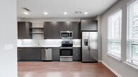 This single-floor residence offers a well-designed layout with modern finishes and a cohesive contemporary aesthetic. The kitchen is a standout feature, showcasing dark flat-front cabinetry extending to the ceiling for generous storage, light quartz countertops, a large stainless steel undermount sink, and a full suite of stainless steel appliances including a French door refrigerator with ice maker, electric range, built-in microwave, and dishwasher. A subtle tile backsplash with a staggered pattern adds texture and visual interest. Recessed lighting illuminates the kitchen and adjoining areas, which flow over durable hardwood-style flooring found throughout the unit. A stylish sliding barn door serves as a functional architectural detail, separating spaces while maintaining an open feel. Built-in open shelving provides practical storage near the entryway. The bedroom is well-proportioned and includes a closet with wire shelving organization systems. The bathroom features a soaking tub with a white subway tile surround, a dark vanity with a light countertop, and wood-look flooring that ties the space together. Natural light filters in through multiple windows fitted with blinds, lending a bright and airy atmosphere to the living areas.