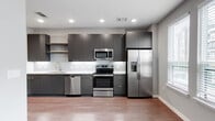 This single-floor residence offers a well-designed layout with modern finishes and a cohesive contemporary aesthetic. The kitchen is a standout feature, showcasing dark flat-front cabinetry extending to the ceiling for generous storage, light quartz countertops, a large stainless steel undermount sink, and a full suite of stainless steel appliances including a French door refrigerator with ice maker, electric range, built-in microwave, and dishwasher. A subtle tile backsplash with a staggered pattern adds texture and visual interest. Recessed lighting illuminates the kitchen and adjoining areas, which flow over durable hardwood-style flooring found throughout the unit. A stylish sliding barn door serves as a functional architectural detail, separating spaces while maintaining an open feel. Built-in open shelving provides practical storage near the entryway. The bedroom is well-proportioned and includes a closet with wire shelving organization systems. The bathroom features a soaking tub with a white subway tile surround, a dark vanity with a light countertop, and wood-look flooring that ties the space together. Natural light filters in through multiple windows fitted with blinds, lending a bright and airy atmosphere to the living areas.