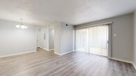 This single-story space presents a clean, modern aesthetic throughout, with a neutral gray and white color palette that creates a bright, cohesive atmosphere. The main living area is generously proportioned, featuring light wood-style plank flooring that extends throughout the primary spaces. A large sliding glass door fitted with vertical blinds serves as the focal point of the living room, allowing natural light to filter in and providing access to an outdoor balcony area.

The layout connects the living area to a hallway leading to the remaining rooms. The kitchen is configured in a galley style, outfitted with white cabinetry, granite-style laminate countertops, a stainless steel sink, and black appliances including a refrigerator, electric range, microwave, and dishwasher. The bathroom continues the updated finishes, featuring a white vanity with a granite countertop, a tiled tub-and-shower surround, and wood-style flooring. The bedroom offers plush carpeting underfoot and a ceiling fan with a light fixture overhead, adding comfort and functionality. Throughout the space, white trim and baseboards provide a crisp contrast to the soft gray walls.