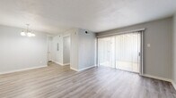 This single-story space presents a clean, modern aesthetic throughout, with a neutral gray and white color palette that creates a bright, cohesive atmosphere. The main living area is generously proportioned, featuring light wood-style plank flooring that extends throughout the primary spaces. A large sliding glass door fitted with vertical blinds serves as the focal point of the living room, allowing natural light to filter in and providing access to an outdoor balcony area.

The layout connects the living area to a hallway leading to the remaining rooms. The kitchen is configured in a galley style, outfitted with white cabinetry, granite-style laminate countertops, a stainless steel sink, and black appliances including a refrigerator, electric range, microwave, and dishwasher. The bathroom continues the updated finishes, featuring a white vanity with a granite countertop, a tiled tub-and-shower surround, and wood-style flooring. The bedroom offers plush carpeting underfoot and a ceiling fan with a light fixture overhead, adding comfort and functionality. Throughout the space, white trim and baseboards provide a crisp contrast to the soft gray walls.
