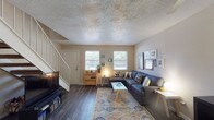 This two-story home offers a well-organized layout designed for comfortable, everyday living. The main level opens into a bright living room featuring dark hardwood-style flooring, a textured ceiling, and multiple windows that allow natural light to fill the space. A staircase with a white railing leads to the upper level, while a front entry door adds accessibility directly from the living area. The kitchen is efficiently arranged in a galley style, equipped with white cabinetry, black appliances including an electric range and refrigerator, and a peninsula counter with a built-in sink — ideal for both cooking and casual use. A full bathroom on this level includes a single-sink vanity with a speckled countertop, a tub-shower combination, and hardwood-style flooring. Upstairs, the spacious primary bedroom features plush carpeting, a ceiling fan, and direct views of the upper landing and staircase. A dedicated home office provides a quiet workspace with a window overlooking greenery, carpet flooring, a ceiling fan, and ample built-in shelving for books and equipment. An additional carpeted room with a large sliding closet offers flexible use as a secondary bedroom or lounge space. Ceiling fans are present throughout the upper level, enhancing comfort across all rooms.