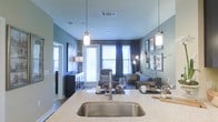 This single-floor residence showcases a thoughtfully designed open-concept layout that seamlessly connects the kitchen and living areas. The kitchen is a standout feature, boasting sleek granite or stone composite countertops with an undermount stainless steel sink, complemented by elegant pendant lighting that hangs from the ceiling to create a warm, focused ambiance. The teal-toned walls lend a cool, contemporary character throughout the space.

The living area is bathed in natural light, courtesy of large floor-to-ceiling windows dressed with both blinds and dark curtain panels, offering flexible light control. The space is well-proportioned, allowing for comfortable furniture arrangements including seating and accent pieces. Wall-mounted artwork and decorative fixtures add personality and depth to the room.

The open sightlines from the kitchen island through to the living area create a sense of expansiveness and connectivity, making the space ideal for both everyday living and entertaining. Recessed ceiling lighting and pendant fixtures work in tandem to provide layered illumination throughout. Overall, the residence combines modern finishes, functional design, and a cohesive color palette to deliver a polished and inviting living environment.