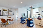 This thoughtfully designed single-floor residence offers a seamless open-concept layout connecting the living, dining, and kitchen areas beneath recessed lighting and smooth white ceilings. Light hardwood-style flooring flows throughout, lending warmth and continuity to the space. The living room is anchored by a striking decorative ceiling fan and floor-to-ceiling glass doors that open to a private balcony, flooding the interior with natural light and offering sweeping city views. The gourmet kitchen is a standout feature, showcasing a large waterfall-edge island with bar seating, sleek two-tone cabinetry in white and charcoal, a textured tile backsplash, stainless steel appliances, a range hood, and globe pendant lighting. An adjacent dining area comfortably accommodates a round table and mid-century modern chairs. The primary bedroom is a serene retreat highlighted by dramatic floor-to-ceiling windows overlooking a lush tree canopy, complemented by contemporary wall sconces and curated artwork. Its en-suite bathroom features a double vanity with illuminated mirrors, charcoal cabinetry, white quartz countertops, and a glass-enclosed walk-in shower. A secondary bedroom and an additional well-appointed bathroom with a walk-in closet complete this refined and cohesive residence.
