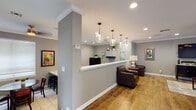 This single-floor space offers a thoughtfully designed layout that blends functionality with a warm, inviting aesthetic. The reception area greets visitors with a sleek front counter framed by a structural column and illuminated by a series of elegant glass pendant lights. Recessed lighting and crown molding add a polished touch, while dark leather armchairs provide a comfortable seating area for guests.

The living room is generously proportioned and anchored by a large sectional leather sofa, a glass-top coffee table, and a wall-mounted flat-screen television atop a dark media console. Natural light streams through wide windows dressed with horizontal blinds and teal drapery panels, complemented by a ceiling fan overhead. A compact dining table with four chairs sits nearby, offering a casual spot for meals or meetings.

The kitchen is efficiently laid out with dark espresso cabinetry, light granite countertops, and black appliances including a range, refrigerator, and range hood. A breakfast bar with industrial-style stools extends into the space, creating a seamless connection to the adjacent dining nook. The nook features tile flooring, corner windows that invite natural light, and colorful framed artwork that adds personality throughout. Crown molding and consistent gray-toned walls unify the entire space with a cohesive, modern feel.