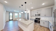 This single-story home offers a thoughtfully designed layout encompassing three bedrooms, two bathrooms, a living room, kitchen, and a dedicated laundry room. The open-concept kitchen serves as the heart of the home, showcasing gray shaker-style cabinetry with black hardware, granite countertops, a mosaic tile backsplash, and a full suite of stainless steel appliances including a refrigerator, range, and microwave. A central island with an undermount sink and pendant globe lighting adds both function and style, flowing seamlessly into the living area. The living room features warm wood-look flooring, a ceiling fan, and multiple windows that invite natural light and outdoor views. The bedrooms are finished with plush carpeting and feature gray panel doors, with at least one bedroom offering a double-door closet. The two bathrooms are finished consistently with granite-topped gray vanities, wood-look flooring, and marble-tile surround tub and shower combinations. One bathroom also includes a separate glass-enclosed shower. The dedicated laundry room accommodates a full-size washer and dryer side by side, complete with wire shelving above. Throughout the home, a neutral color palette and cohesive finishes create a clean, modern aesthetic.
