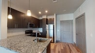 This single-floor unit showcases a contemporary design with a cohesive, modern aesthetic throughout. The kitchen serves as a central highlight, featuring dark espresso-toned cabinetry that extends to the ceiling, a white subway tile backsplash, and granite countertops. Stainless steel appliances — including a refrigerator, range, and built-in microwave — contribute to a polished, functional workspace. A peninsula with an undermount sink and pendant lighting above adds both style and additional prep space, seamlessly connecting the kitchen to the open living and dining area.

The living area is finished with wood-style plank flooring in warm, varied tones, creating a bright and welcoming atmosphere. Neutral walls complement the flooring and allow for versatile décor options. The bedroom features plush carpeting and a large window fitted with horizontal blinds, allowing ample natural light while maintaining privacy and offering views of the surrounding area.

The bathroom continues the unit's design language with granite countertops, an undermount sink, chrome fixtures, and a large frameless mirror. The overall layout, visible in the floor plan view, is efficiently organized, with clear separation between the living, sleeping, and bathroom areas. The unit presents a clean, move-in-ready aesthetic suited for modern living.