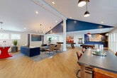 Located at 22 Country Club Dr in Manchester, NH, this single-floor amenity space offers an impressive and well-appointed layout designed for recreation and fitness. The open-concept game room serves as the centerpiece, featuring soaring vaulted ceilings with a bold navy blue accent, warm hardwood-style flooring, and an abundance of natural light streaming through a generous row of windows. A large-screen TV anchors a comfortable lounge area with a navy sectional sofa and leather accent chairs, while a dedicated billiards area complete with a pool table and red pendant lighting adds to the entertainment appeal. A wet bar with white cabinetry and a striking navy tile backsplash provides a functional refreshment station. Decorative touches, including an American flag wall art piece and blueprint-style artwork, lend the space a distinct, curated character. French doors in the game room open to an outdoor deck, extending the entertaining area further. The fully equipped gym occupies a spacious separate area, outfitted with a comprehensive array of Cybex cardio machines, weight stations, cable systems, free weights, and cycling bikes. Bold red and white striped wall detailing and a red, white, and blue floor accent give the gym an energetic, athletic atmosphere. Two bathrooms and a closet round out the functional amenities of this versatile space.