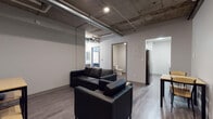 Located in the heart of downtown Denver at 1031 14th St, this single-floor unit blends industrial aesthetics with functional modern design. The living area is defined by striking exposed concrete ceilings with visible ductwork and piping overhead, lending the space a raw, urban character. Durable wood-look flooring runs throughout, tying together the open layout. The lounge is furnished with black leather seating arranged around a compact coffee table, creating a practical gathering space. An open shelving unit provides additional storage and display space within the lounge area. The sleeping quarters offer a private retreat, each featuring a bed with under-frame storage and a wood-and-steel wardrobe unit for clothing and personal items. Natural light filters in through a window, brightening the room. The galley-style kitchen is efficiently laid out with white upper cabinetry, ample counter space with a clean white surface, a tiled backsplash, and black stainless appliances including a range and microwave. The bathroom is clean and straightforward, featuring a walk-in shower stall, toilet, and wood-look vinyl flooring consistent with the rest of the unit. Overall, the space delivers a utilitarian yet contemporary urban living environment well-suited to downtown Denver.